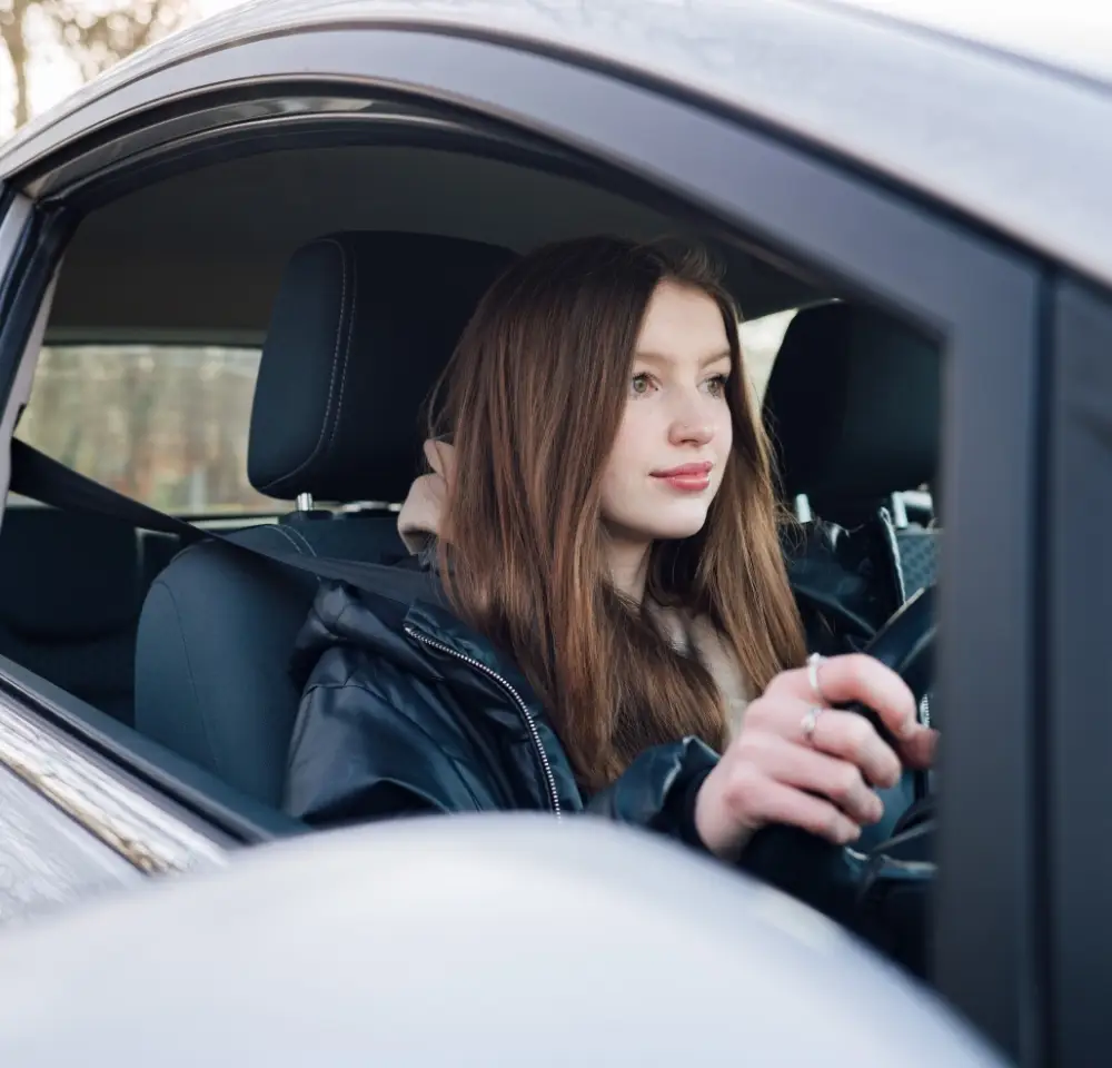 A young woman with long brown hair sits in the driver’s seat of a car, looking forward with both hands on the steering wheel, wearing a dark jacket and seatbelt—ready to hit the road with her old car insurance.