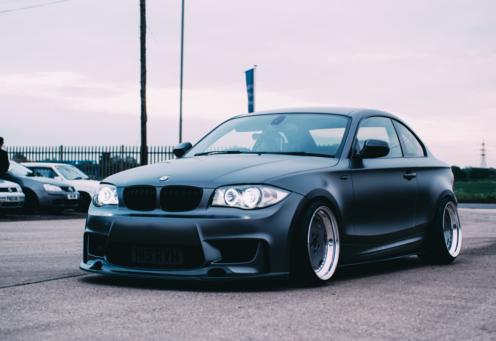 A matt black BMW coupé with custom wide-body kit and lowered suspension is parked outdoors on a cloudy day, perfect for showcasing its unique style and worth—an ideal candidate for Agreed Value Car Insurance. Other cars and a fence appear in the background.