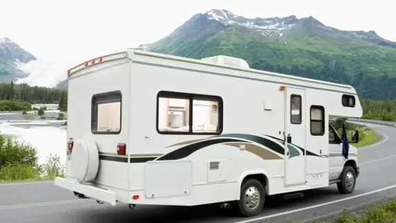 A white campervan drives along a winding road with green mountains and trees in the background. The windows of the campervan are illuminated, and a river flows nearby under a cloudy sky.