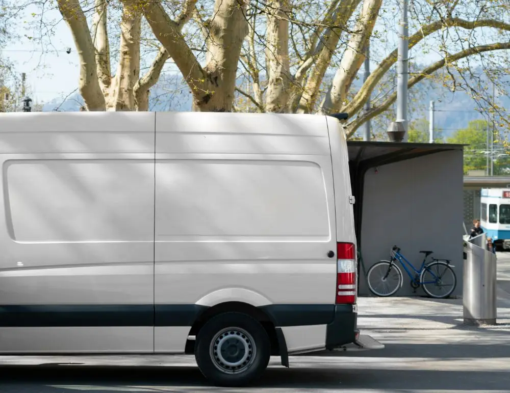 A white delivery van, covered by Mercedes Van Insurance, is parked on a city street near a grey bike shelter with a bicycle. Leafy trees stretch overhead and a tram is visible in the background.
