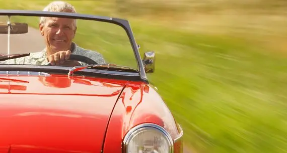 An older man smiles whilst driving a red convertible car with the top down on a sunny day, with a blurred green landscape in the background suggesting movement.