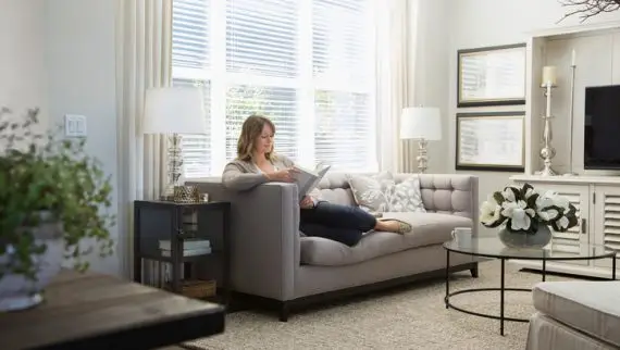 A woman sits on a light grey sofa reading a book in a bright, modern living room with large windows, white curtains, lamps, framed pictures, and a glass coffee table with a flower arrangement.