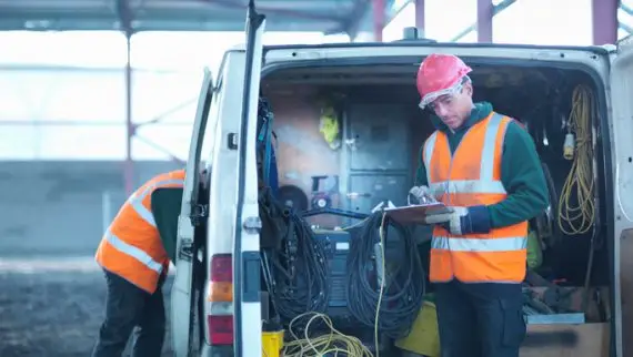 A construction worker in a hard hat and high-visibility vest writes on a clipboard beside an open work van, whilst another worker searches inside the van. Tools and cables are visible in the van.