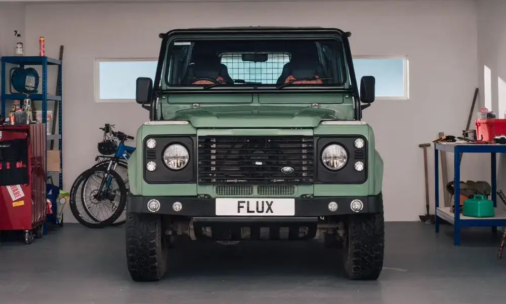A green Land Rover Defender with a FLUX number plate is parked inside a garage, flanked by shelves with tools and a bicycle.