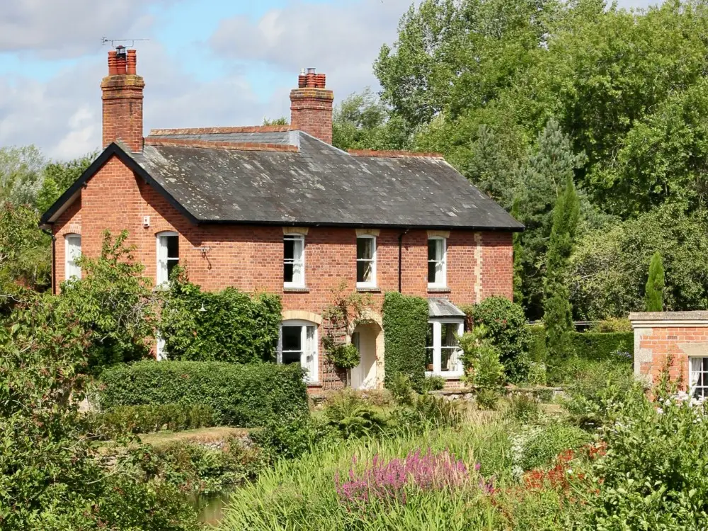 A two-storey red brick house with white trim, surrounded by lush greenery and gardens, stands under a partly cloudy sky with trees in the background. Ivy climbs parts of the house, and flowers bloom in the garden.