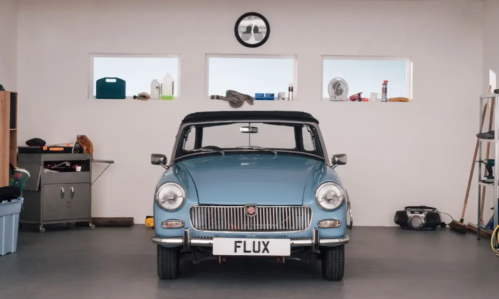 A light blue classic convertible car with a FLUX number plate is parked in a clean, organised garage with white walls, three small windows, and various items on shelves and the floor.