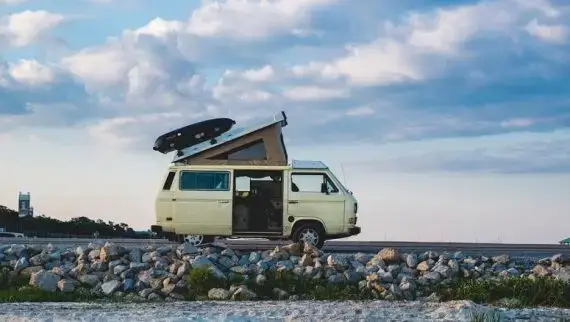 A beige camper van with a pop-up roof and a surfboard on top is parked on a rocky shoreline under a blue sky with scattered clouds.