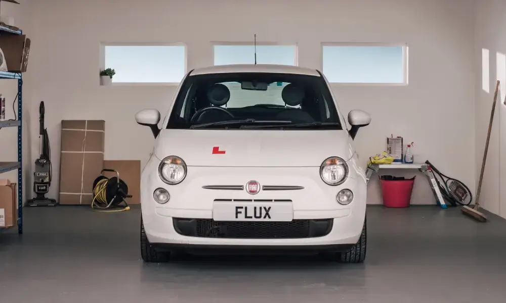 A white Fiat 500 with a learner driver L plate and FLUX number plate is parked in a clean garage with shelves, boxes, a vacuum cleaner, and sports equipment visible around it.