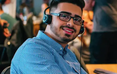 A man wearing glasses and a blue striped shirt smiles whilst working at a desk, wearing a headset in a busy office environment.