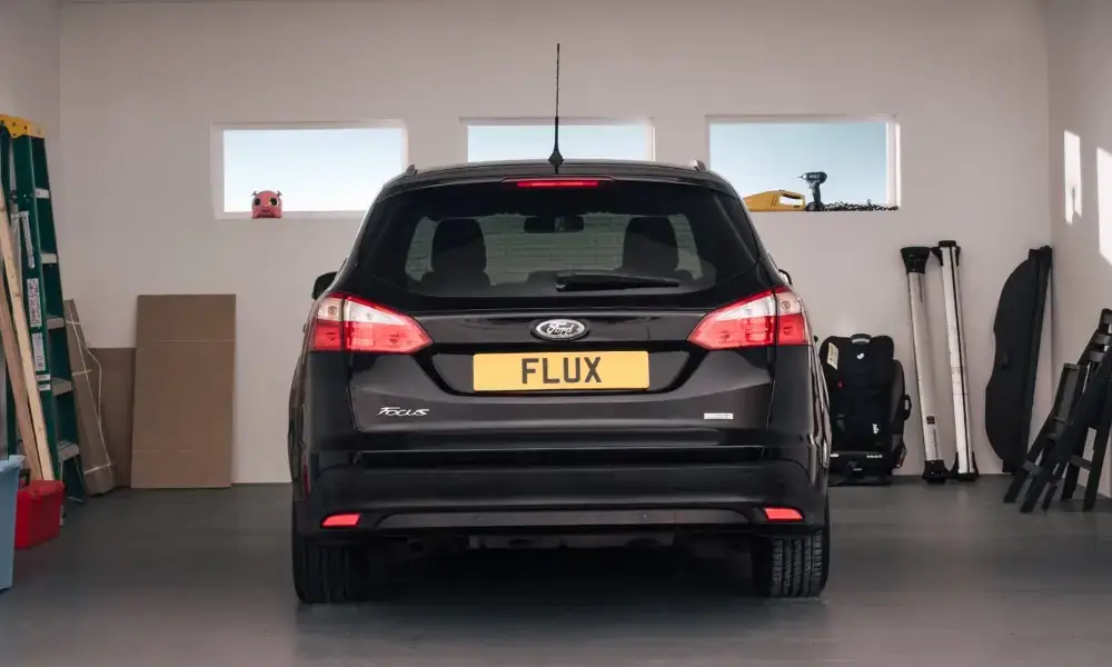 A black Ford Focus with a yellow number plate reading FLUX is parked inside a clean garage with tools, boxes, and folded equipment along the walls.