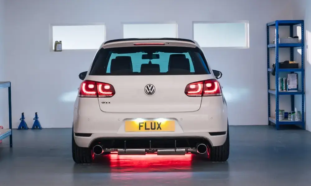 A white Volkswagen GTI is parked in a clean, well-lit garage. The rear lights are on, a custom exhaust and red underglow are visible, and the number plate reads FLUX. Shelving and tools are seen in the background.