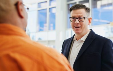 A man in a suit and glasses smiles whilst talking to another person in an orange shirt in a well-lit indoor setting.
