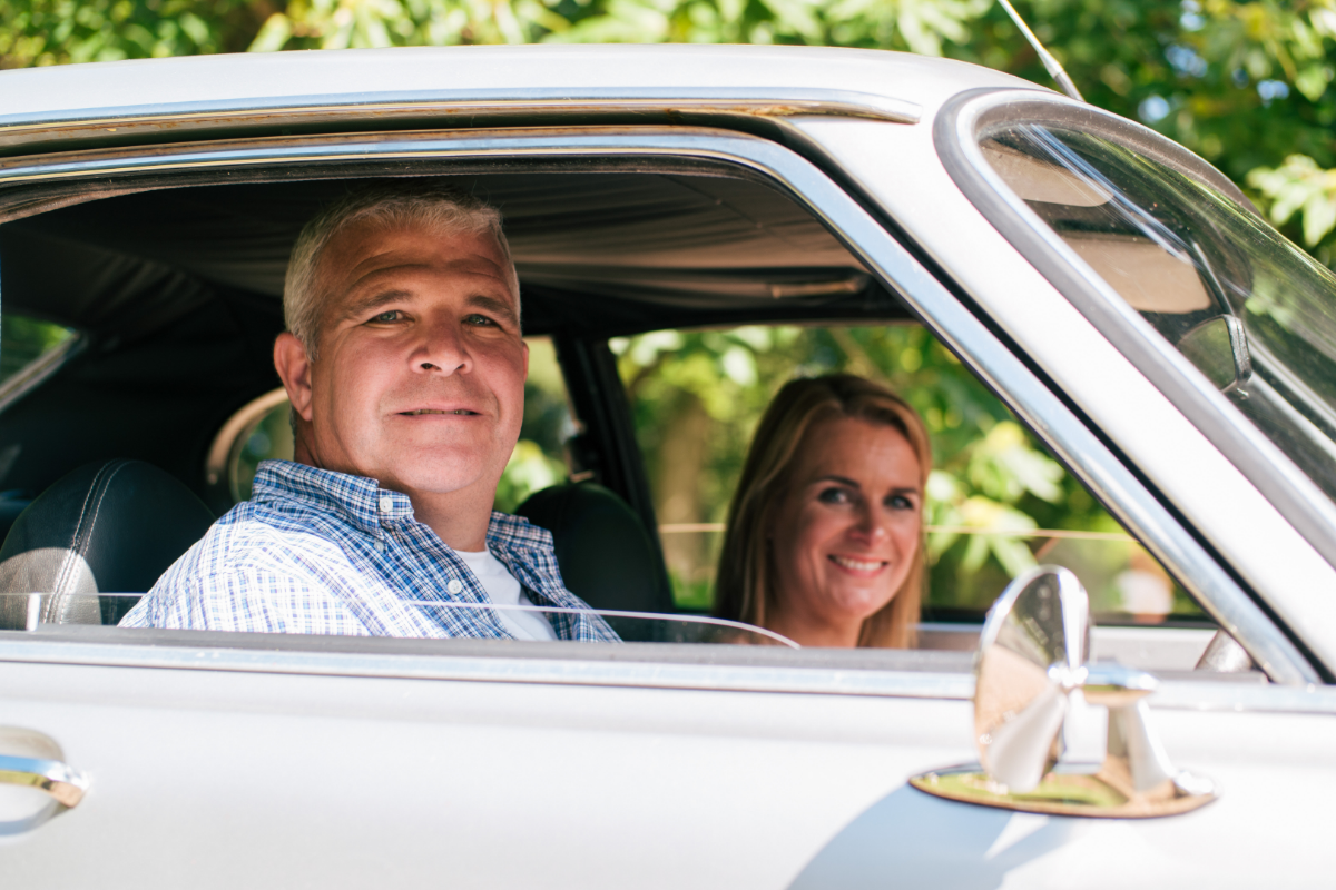 A man and a woman sit inside a car with the windows down, smiling at the camera—perfectly enjoying their drive with Individual Insurance. Sunlight filters through green trees in the background.